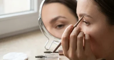 Mano definiendo cejas de forma natural con un lápiz mecánico de punta fina frente a un espejo, con un cepillo de carrete sobre el tocador